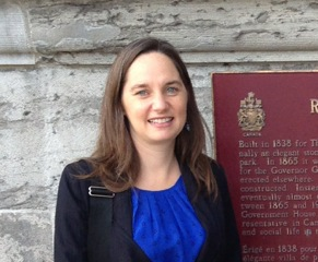 Picture of Eryka, in front of a stone wall and a red historical plaque. She wears a black blazer over blue blouse and is smiling. Erika Dyck 
